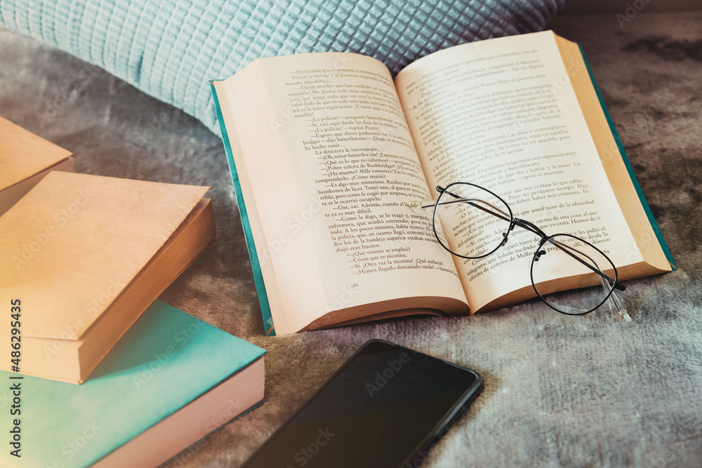 Obraz premium Medium shot. Glasses resting on the open book, next to the cushion, the mobile phone and some books on the bed in the bedroom in the morning.