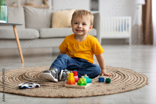 Cute toddler boy playing with car and educational wooden toy at home, sitting on floor carpet in living room and smiling
