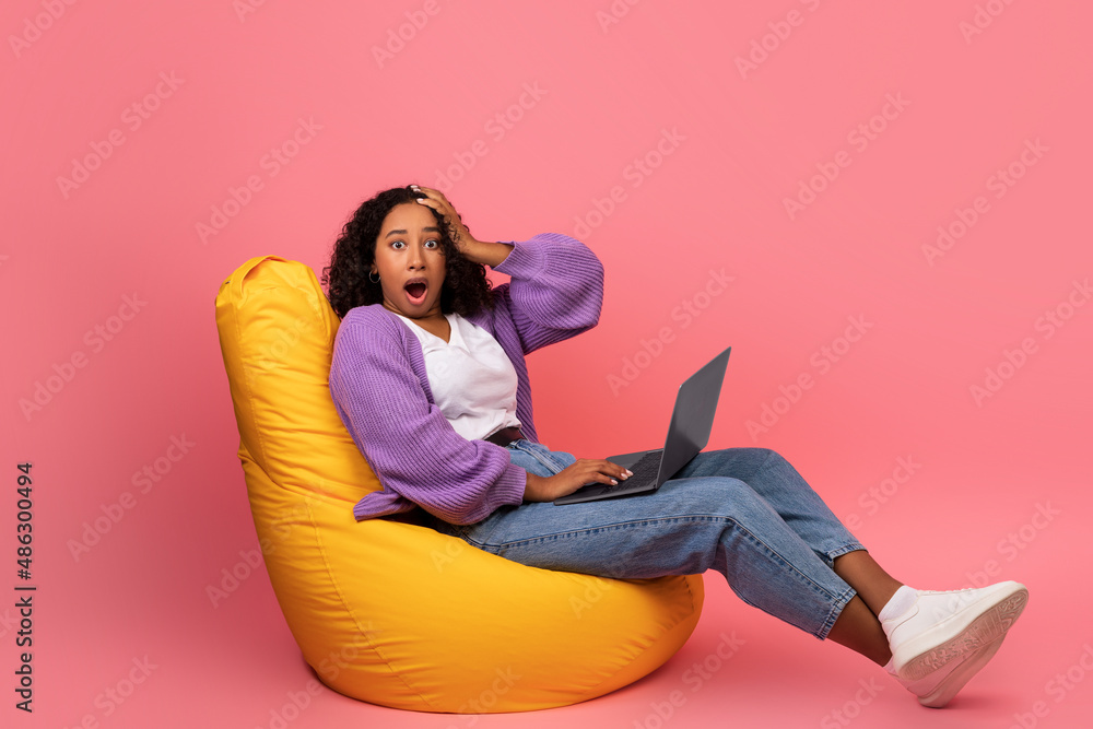 Portrait of shocked young black woman sitting in bean bag chair with laptop computer and grabbing her head in terror