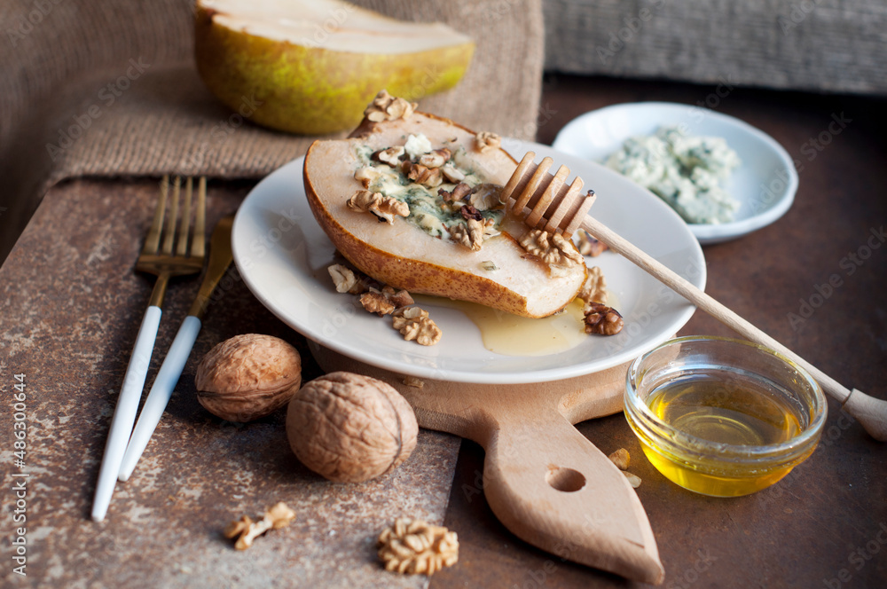 Pear baked with blue cheese, honey and walnuts on a white plate on a brown and wooden background. Backed pear with fork, knife and wooden honey stick. Rustic style Pear baked with blue cheese, honey and walnuts on a white plate on a brown and wooden background. Backed pear with fork, knife and wooden honey stick. Rustic style