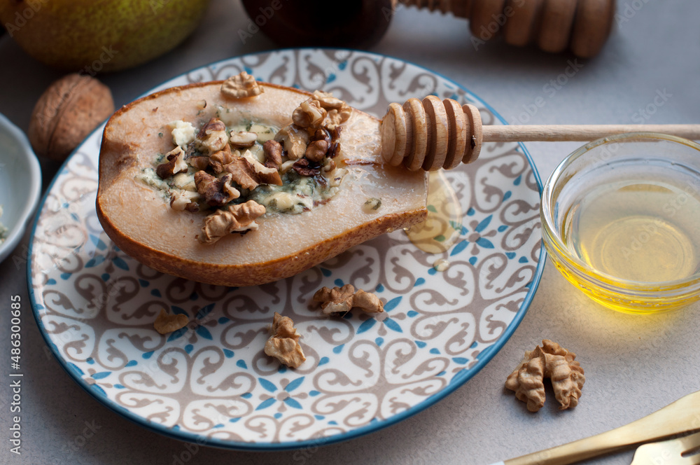 Pear baked with dorblu cheese, honey and walnuts on a plate on a gray background. Backed pear with wooden honey stick Pear baked with dorblu cheese, honey and walnuts on a plate on a gray background. Backed pear with wooden honey stick