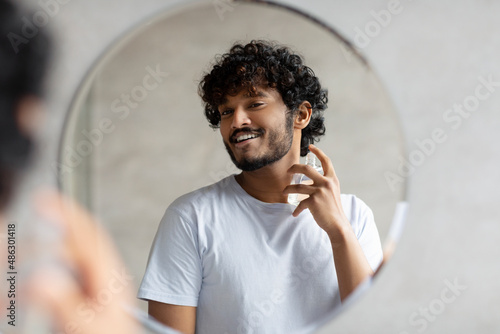 Photos Happy indian man looking in mirror spraying on cologne in bathroom interior, doi