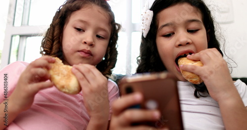 Generation Z little girls using mobile phone while eating sandwich snack