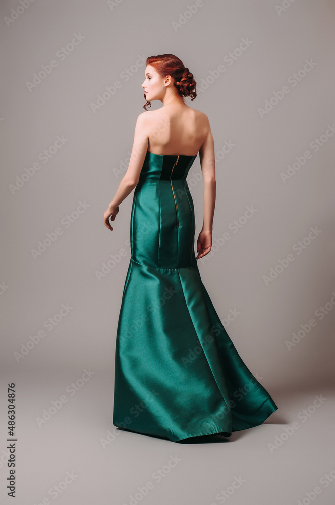 Emerald ball gown. Young lady in long green evening dress. Ginger woman posing in studio. Beautiful model wearing high heels, feminine look for an event. Women's classic fashion.