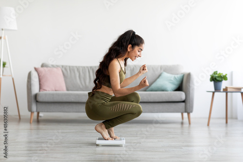 Triumphant millennial Indian woman sitting on scales, making YES gesture, excited about her weight loss result at home