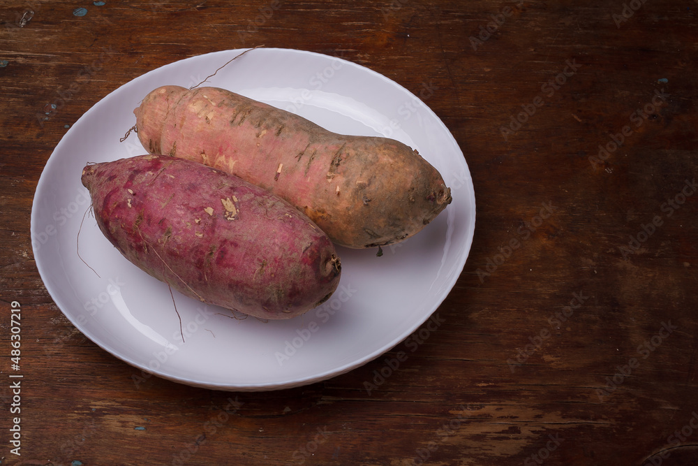 Orange, purple raw sweet potatoes, yam on white plate, wooden rustic table. Farm harvest, organic vegetables, shop local vegan food Orange, purple raw sweet potatoes, yam on white plate, wooden rustic table. Farm harvest, organic vegetables, shop local vegan food