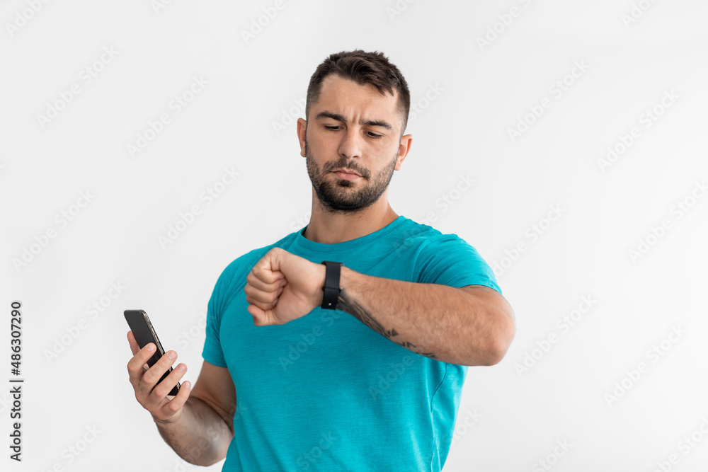 Serious young caucasian man looking at fitness tracker and hold smartphone, check pulse and sport result Serious young caucasian man looking at fitness tracker and hold smartphone, check pulse and sport result