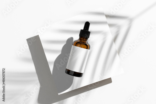 Top view of brown dropper glass bottle lies on white podium. Skincare concept. Mock-Up organic natural cosmetic product. Light background with palm leaf shadow. Minimalistic showcase photo