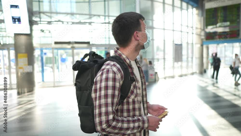 Travel during coronavirus quarantine restrictions. Man in medical mask from infection, viruses or allergies takes out passport and boarding pass from back pocket in terminal near board of departures. Travel during coronavirus quarantine restrictions. Man in medical mask from infection, viruses or allergies takes out passport and boarding pass from back pocket in terminal near board of departures.