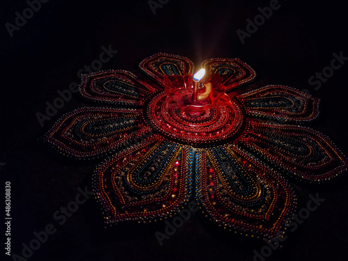 Stock photo of beautiful red color plastic oil lamp lit especially during diwali festival kept on colorful rangoli under dark background. Picture captured at Bangalore, Karnataka, India. focus on lamp photo