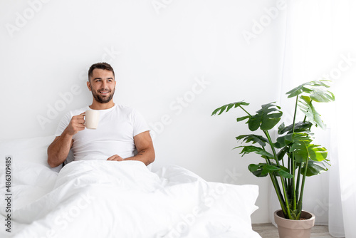 Happy glad millennial caucasian handsome muscular male sits on white bed, drinks coffee, enjoy good morning #486309485