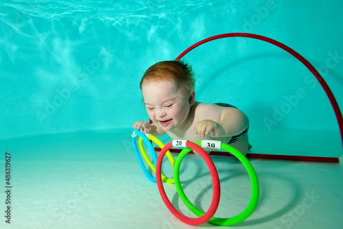 A happy kid with Down syndrome swims and collects toys underwater from the bottom of a children's pool during a sports activity. Children's disability. Concept. Portrait. Horizontal orientation.