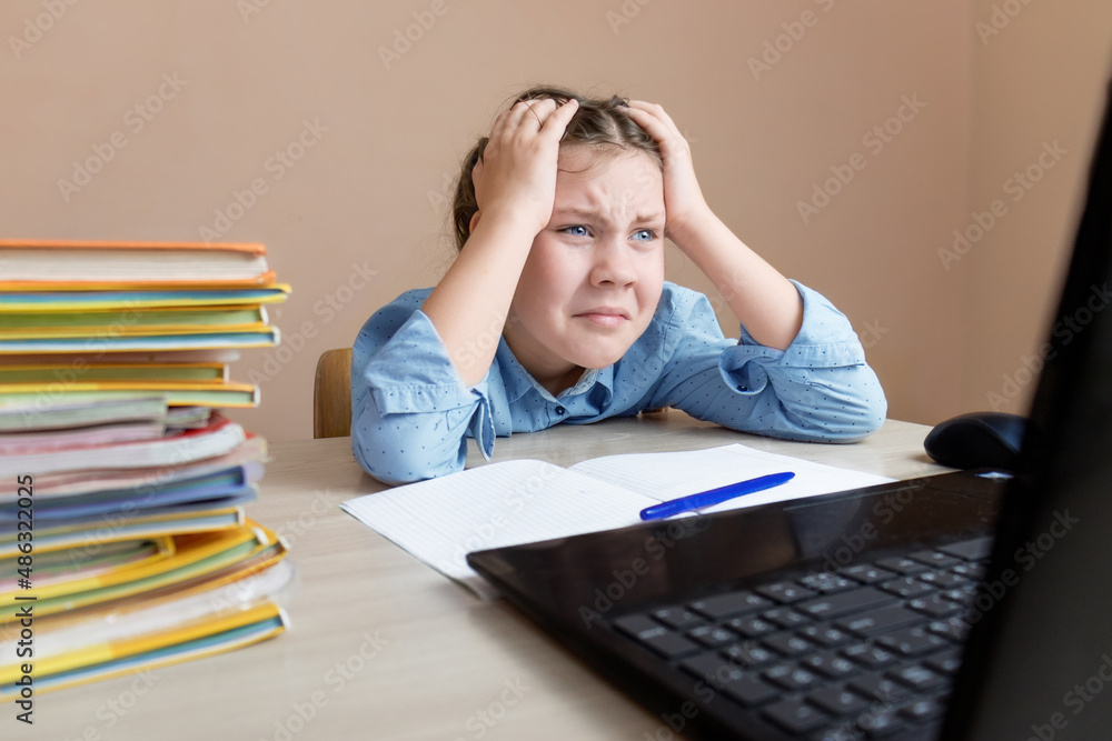 girl child sits at the table and does online lessons and looks with fatigue and misunderstanding and despair online lesson by laptop.online education, stress girl child sits at the table and does online lessons and looks with fatigue and misunderstanding and despair online lesson by laptop.online education, stress
