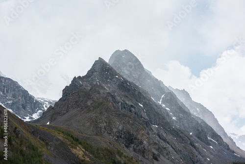 Atmospheric mountain landscape with large pointy pinnacle under gray cloudy sky. Gloomy scenery with high pointed mountain peak with sharp rocks in low clouds. Huge rocky peaked top in rainy weather.