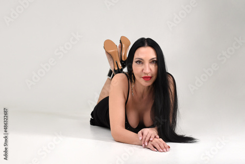 A brunette girl beautiful and stylish in a black dress lies belly down with her hand under her chin in the studio in an attractive pose on a white background photo