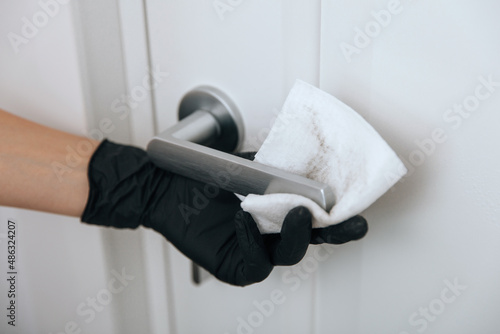 Cleaning door handle with dirty wipe in black gloves. Sanitize surfaces prevention in hospital and public spaces against corona virus. Woman hand using towel for cleaning home room door link