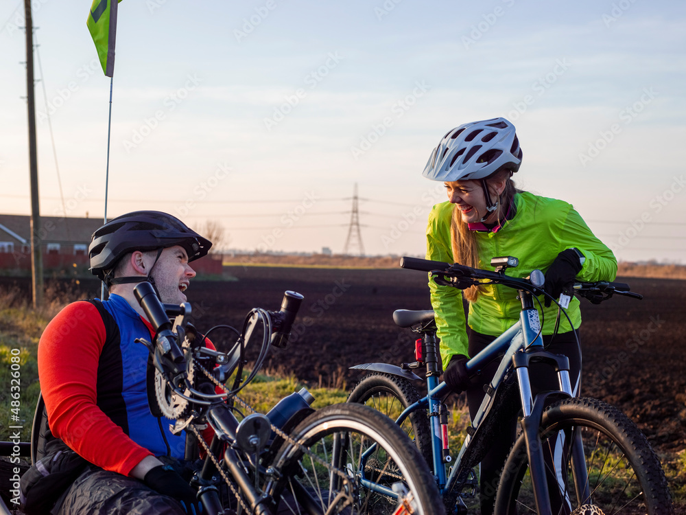 Disabled man on handcycle and woman with bicycle laughing in rural landscape