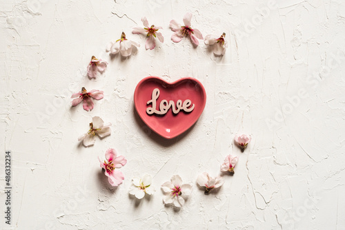 White textured Valentine's background with circle of almond blossoms and a pink heart-shaped plate with words written on wood 