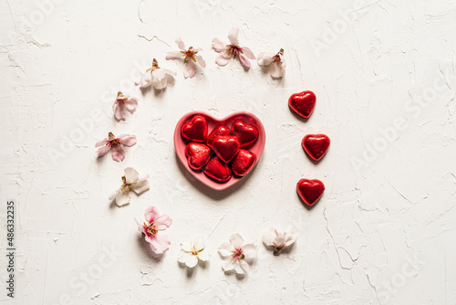 White textured Valentine's background with circle of almond blossoms and a pink heart-shaped plate with red heart-shaped chocolates. Romantic flat lay