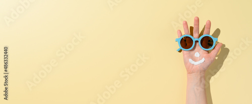 Funny smiling face created from sunscreen cream and sunglasses on female hand. Minimal summer scene on sandy-colored background. Health care and beauty concept. Copy space, banner