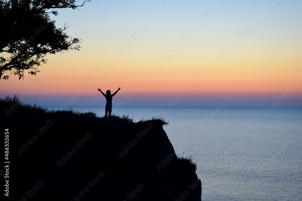 Silhouette of an unrecognizable woman with outstretched arms near the branches of a tree against the backdrop of the evening sea