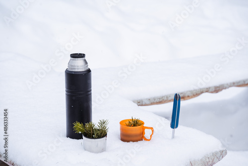 A black thermos and mugs with el's sprig in the snow. A hiking knife will be stuck in the snow. Winter still life in the snow photo