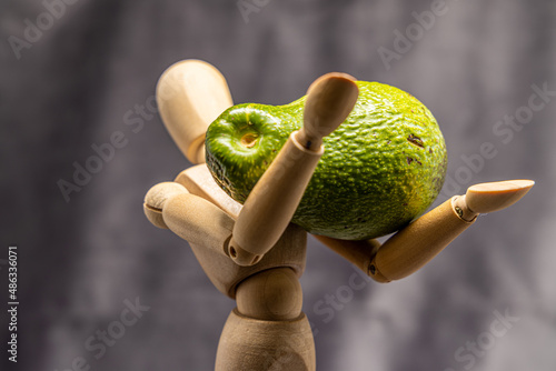 A closeup shot of a small wooden human mannequin holding a ripe avocado against a gray background