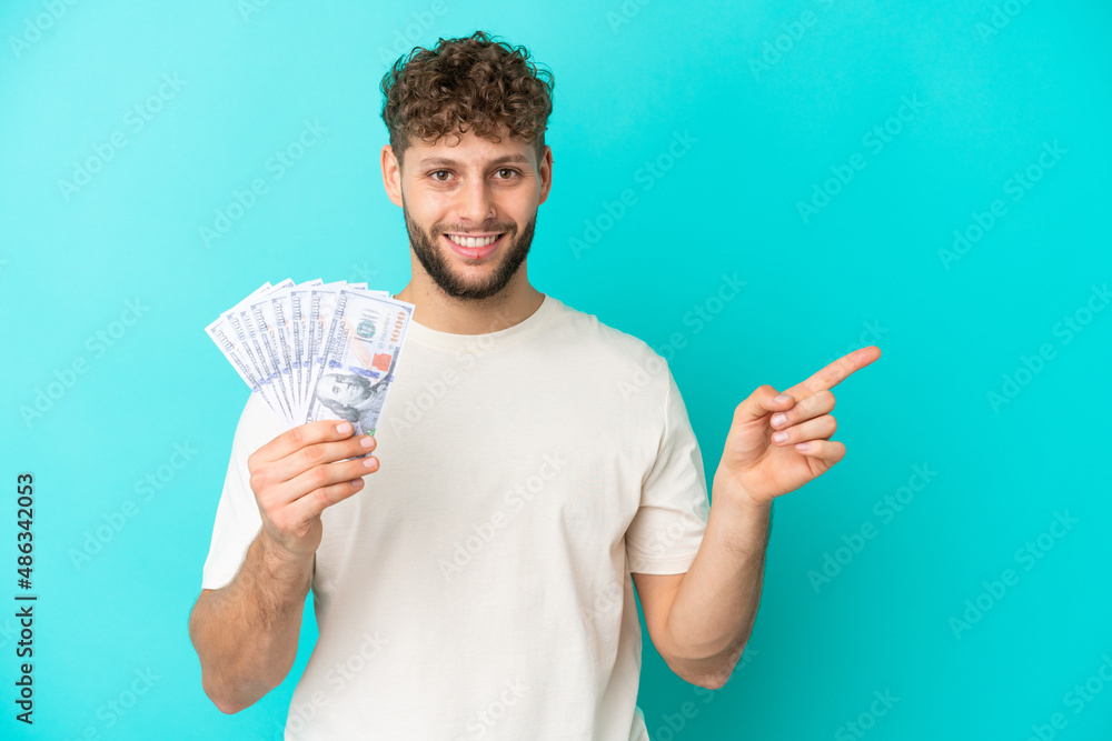 Young caucasian man taking a lot of money isolated on blue background pointing finger to the side Young caucasian man taking a lot of money isolated on blue background pointing finger to the side