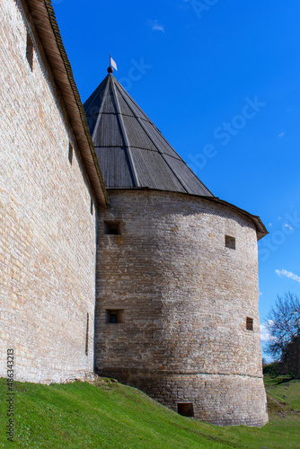 Round fortress tower with loopholes at the junction with the fortress wall. A tower with a hipped wooden roof. Side view. Early spring. Daylight. Clear sky.