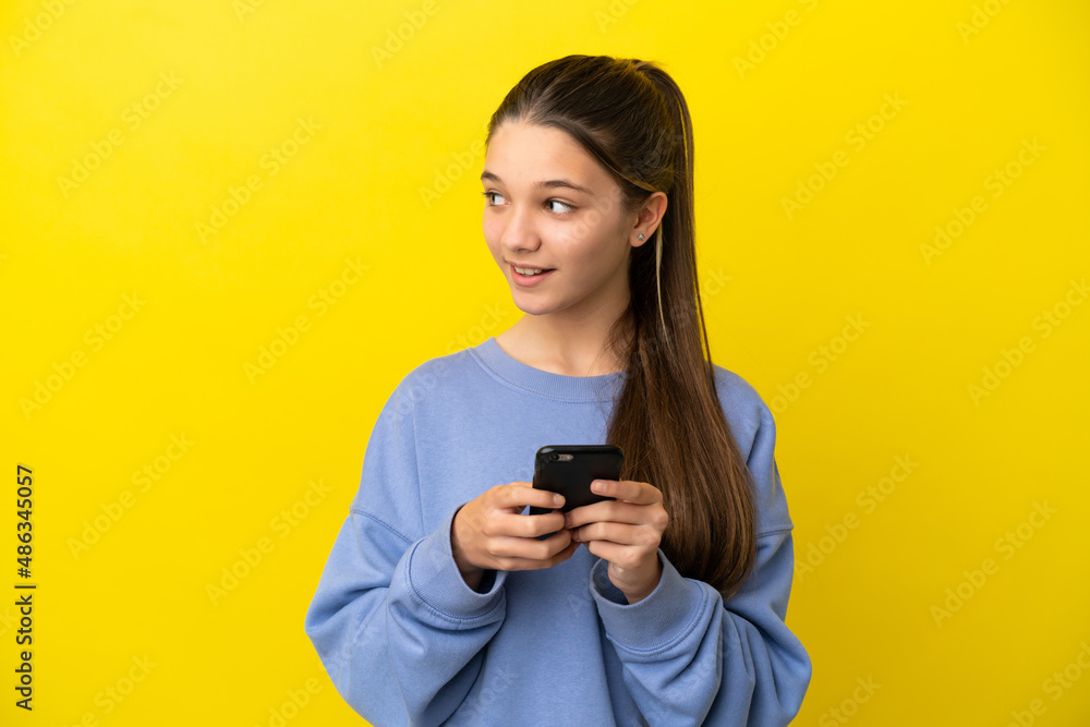 Little girl over isolated yellow background using mobile phone and looking up Little girl over isolated yellow background using mobile phone and looking up