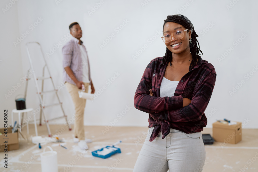 Proud woman in glasses mother of two daughters stands and smiles to camera with arms crossed in background husband is painting walls of new apartment, girls come to help in the renovation Proud woman in glasses mother of two daughters stands and smiles to camera with arms crossed in background husband is painting walls of new apartment, girls come to help in the renovation