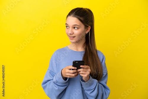 Little girl over isolated yellow background using mobile phone and looking up