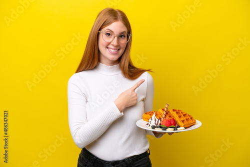 Young redhead woman holding waffles isolated on yellow background pointing to the side to present a product