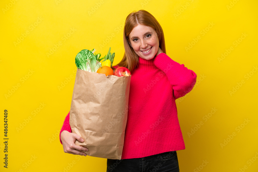 Young redhead woman holding a grocery shopping bag isolated on yellow background laughing Young redhead woman holding a grocery shopping bag isolated on yellow background laughing