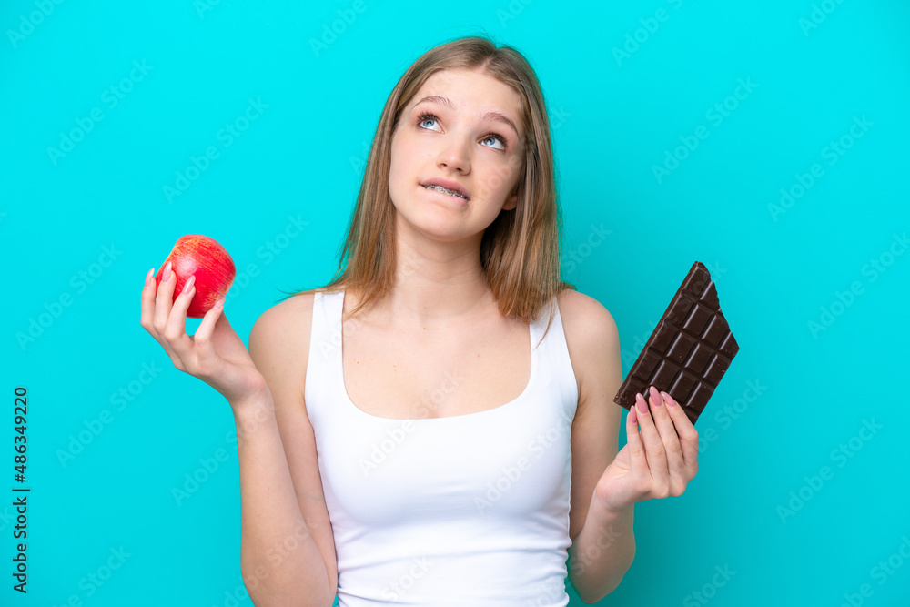 Teenager Russian girl isolated on blue background having doubts while taking a chocolate tablet in one hand and an apple in the other Teenager Russian girl isolated on blue background having doubts while taking a chocolate tablet in one hand and an apple in the other