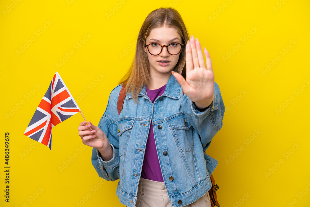 Teenager Russian girl holding an United Kingdom flag isolated on yellow background making stop gesture Teenager Russian girl holding an United Kingdom flag isolated on yellow background making stop gesture