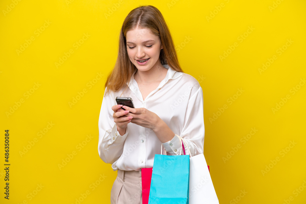 Teenager Russian girl isolated on yellow background holding shopping bags and writing a message with her cell phone to a friend Teenager Russian girl isolated on yellow background holding shopping bags and writing a message with her cell phone to a friend