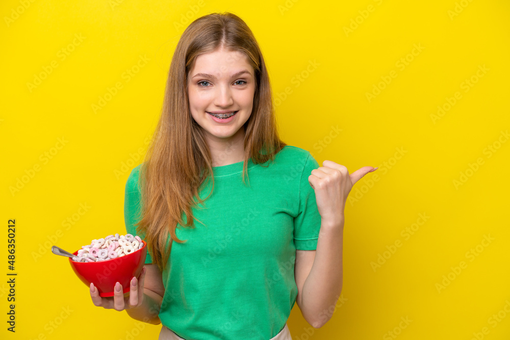 Teenager Russian girl holding bowl of cereals isolated on yellow background pointing to the side to present a product Teenager Russian girl holding bowl of cereals isolated on yellow background pointing to the side to present a product