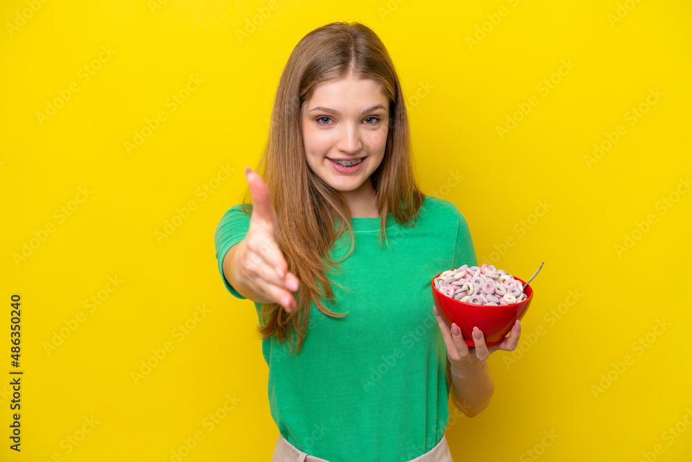 Teenager Russian girl holding bowl of cereals isolated on yellow background shaking hands for closing a good deal Teenager Russian girl holding bowl of cereals isolated on yellow background shaking hands for closing a good deal