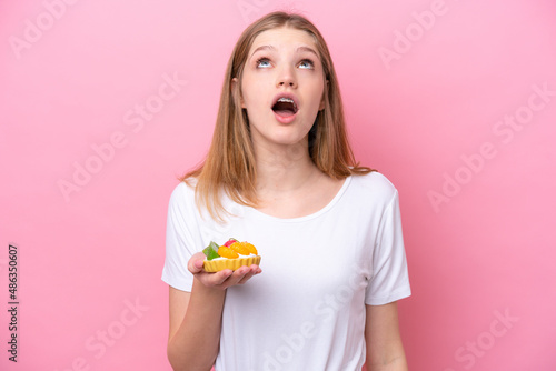 Teenager Russian girl holding a tartlet isolated on pink background looking up and with surprised expression