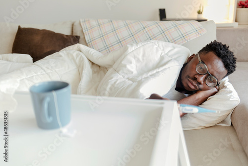 Ill black guy holding thermometer having influenza symptoms and headache lying in bed, indoors. Covid-19 virus, seasonal health problems. Fever And Flu. Healthcare and medicine concept.