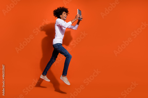 Photo of pretty excited guy dressed white shirt jumping high reading modern gadget empty space isolated orange color background