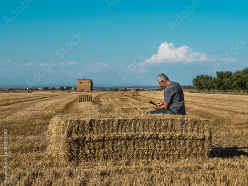 Senior farmer sitting on straw bale looking at mobile phone in field.