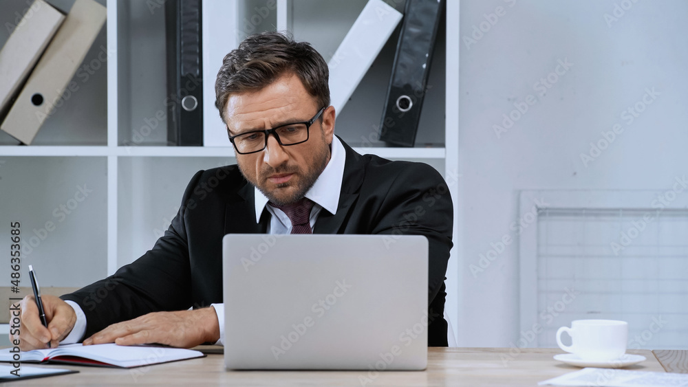 businessman in eyeglasses writing in notebook while looking at laptop in office businessman in eyeglasses writing in notebook while looking at laptop in office
