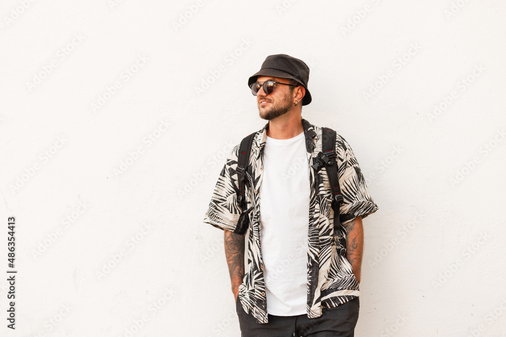 Hipster man in fashion summer clothes with backpack stands near white wall. Guy travel on the street Hipster man in fashion summer clothes with backpack stands near white wall. Guy travel on the street