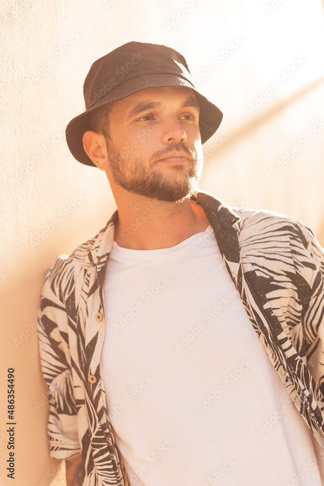 Stylish handsome hipster man with a black panama bucket hat in a fashion beach shirt with palm trees on the beach at sunset Stylish handsome hipster man with a black panama bucket hat in a fashion beach shirt with palm trees on the beach at sunset