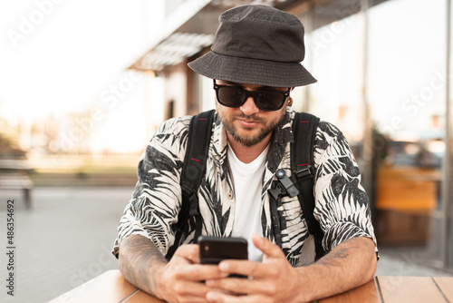 Tableau sur toile Handsome young hipster man in fashionable clothes with a hat sits in a summer cafe and communicates, surfs on a smartphone