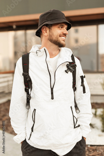 Happy young stylish hipster man in fashion hat with trendy white jacket and backpack travels in the city