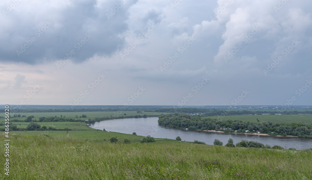 Summer landscape with meadow grass in the foreground. Dark sky with clouds before rain. Bright green grass. Wide river in the background.