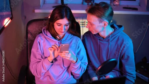 Cute girl and guy in hoodies are talking and fun on camera. Two streamers play video games and interact with the audience. Gamers are sitting in hoodies, chatting on a webcam on a blue-pink background photo